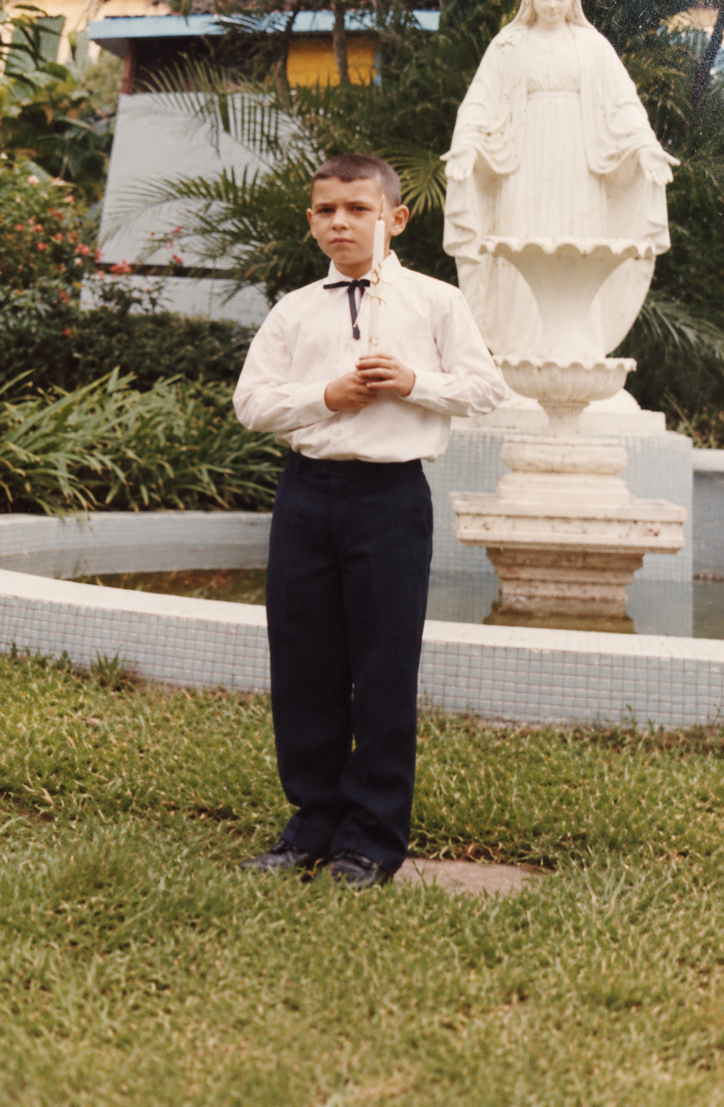 Fotografia antiga de um menino em pé no gramado, junto a uma escultura sobre um pedestal, com um edifício ao fundo.